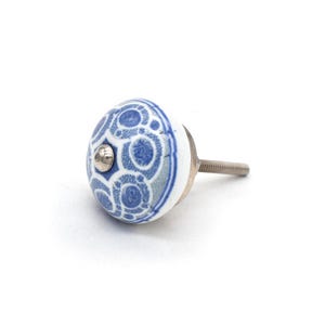 May include: A white and blue ceramic cabinet knob with a decorative pattern. The knob has a silver screw and a small silver accent in the center. The knob is approximately 1.5 inches in diameter.