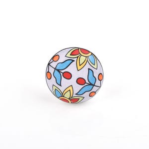 Peut inclure: Un bouton de meuble rond avec un fond gris clair. Le bouton présente un motif floral avec des feuilles bleues, des baies rouges et des fleurs jaunes et rouges. Le bouton est probablement en céramique ou dans un matériau similaire.