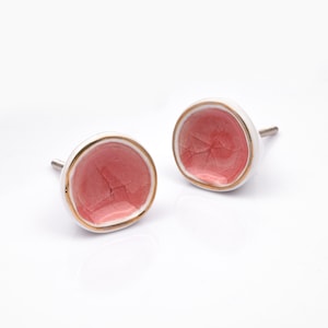 Può includere: Due pomelli rotondi in ceramica per mobili con un centro concavo color rosa polvere e una finitura smaltata craquelé visibile. Ogni pomello presenta un bordo esterno bianco liscio e un sottile profilo metallico dorato, con una vite filettata.