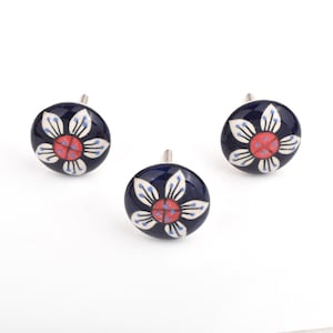 May include: Three navy blue ceramic cabinet knobs with a floral design. Each knob features a white flower with red and blue accents. The knobs have a silver metal stem for mounting and are set against a white background.