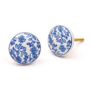 May include: Two round ceramic knobs featuring a white base with a blue floral and vine pattern. Each knob has a smooth, glossy finish and a gold-toned metal screw extending from the back of one piece, designed for attachment to furniture or cabinetry.