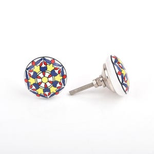 Può includere: Due pomelli decorativi in ceramica per mobili. I pomelli sono bianchi con un colorato motivo floreale in giallo, rosso e blu. Ogni pomello ha una vite metallica per l'installazione. I pomelli sono su uno sfondo bianco.
