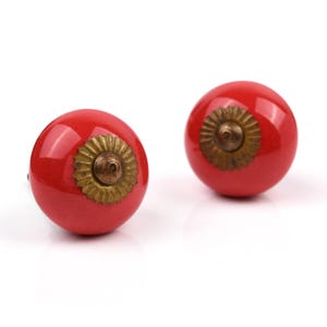 Op de afbeelding: Twee ronde, glanzend rode kastknoppen met een glad, bolvormig oppervlak. Elke knop is voorzien van een decoratieve, geschulpte, messingkleurige metalen plaat in het midden, die een kleine, puntige, messingkleurige verhoging omsluit.