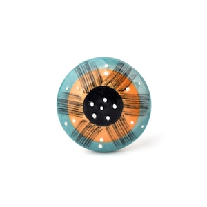 May include: A round ceramic knob with a distinctive pattern. The knob has a teal outer ring, an orange central section, and a black centre. Black lines extend from the centre, and white dots are dotted around the teal ring. A stylish furniture accent.