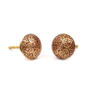 May include: Two decorative cabinet knobs. Each knob is round with a maroon base and a gold floral pattern. The knobs have a gold-colored screw for attachment and are set against a white background.