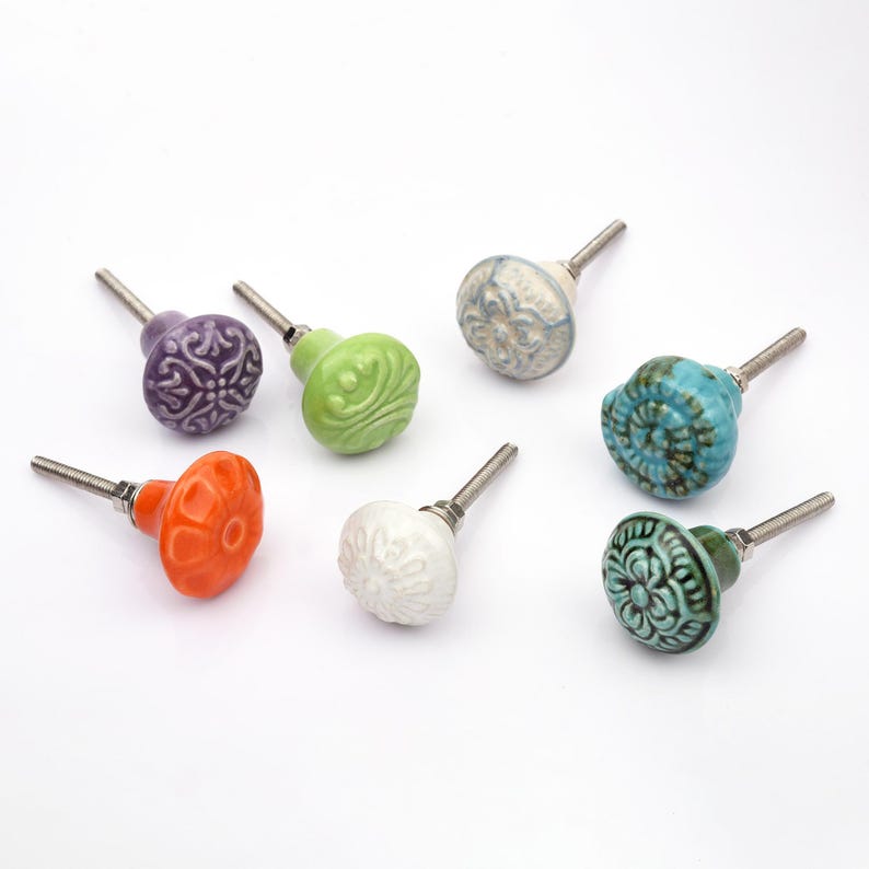 May include: A collection of twelve decorative cabinet knobs in various colours and patterns. Each knob features a round or oval shape with a metal bolt extending from the back. The designs include floral, geometric, and solid textures in shades of green, blue, orange, purple, white, and grey.