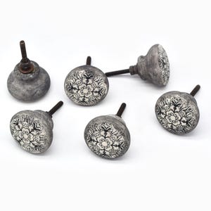 May include: Six grey ceramic cabinet knobs with a black and white floral design. Each knob has a metal screw for installation. The knobs are round and have a distressed, vintage appearance, suitable for furniture or decor.
