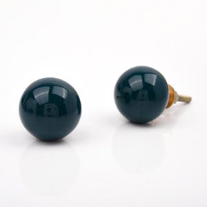 Puede incluir: Dos pomos esféricos para armarios con un acabado liso y brillante en color verde azulado oscuro. Cada pomo presenta una cabeza redonda en forma de bola y un tornillo de montaje metálico que sobresale de la parte posterior.