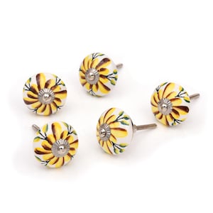 Puede incluir: Un juego de cuatro pomos redondos de cerámica para armarios con un diseño de girasol pintado a mano en amarillo y marrón sobre fondo blanco. Cada pomo tiene un centro de metal plateado y un perno metálico roscado en la parte posterior.
