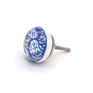 Pode incluir: Um puxador de arm&aacute;rio de cer&acirc;mica branca e azul com um padr&atilde;o floral decorativo. O puxador tem uma base de metal prateado e uma haste roscada para instala&ccedil;&atilde;o. O puxador tem aproximadamente 3,8 cm de di&acirc;metro.