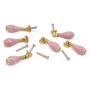 May include: Six pink ceramic cabinet knobs with gold metal bases and screws. The knobs are teardrop shaped and have a speckled finish.