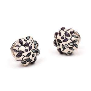 May include: Two round ceramic cabinet knobs with a scalloped edge. The knobs feature an off-white base color with a hand-painted pattern of dark purple and teal leaves and vines outlined in black. Each knob has a silver-toned metal base attachment.