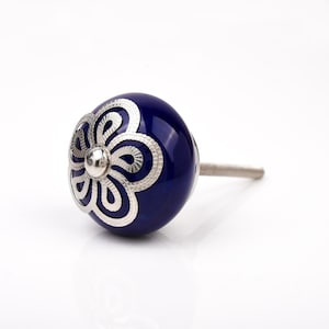 Puede incluir: Pomo de armario de cerámica azul marino con un diseño floral plateado. El pomo tiene forma redondeada y un vástago metálico para la fijación. El diseño plateado está centrado en la cara del pomo.