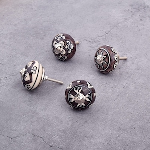 Op de afbeelding: Vier keramische kastknoppen met ingewikkelde bloem- en geometrische patronen in bruin en wit. De knoppen hebben een zilveren metalen schroefbevestiging.