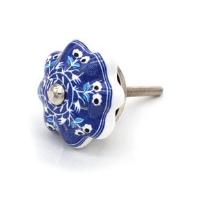 May include: A decorative cabinet knob with a floral design. The knob is a starburst shape with a white scalloped edge and a dark blue center. The center features a white floral pattern and a silver metal screw.