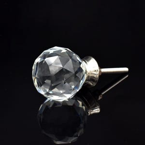 May include: A clear, faceted crystal knob with a silver-toned base and screw. The knob is spherical and reflects light, creating a sparkling effect. The background is black, highlighting the crystal's clarity. Ideal for furniture or cabinet hardware.