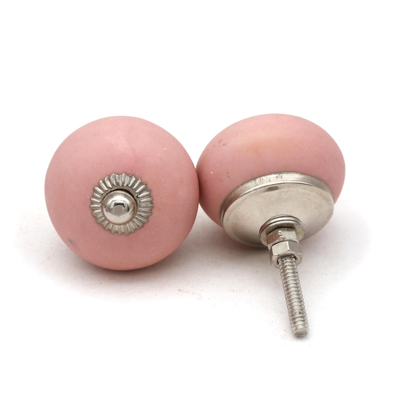 May include: Two pink ceramic cabinet knobs with silver-colored hardware. The knobs are round with a matte finish and a decorative silver accent. The hardware includes a threaded bolt for easy installation.