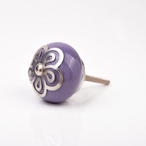 Puede incluir: Un pomo de cerámica lavanda con un diseño floral plateado. El pomo tiene forma redondeada y un vástago metálico roscado para su fijación. El diseño plateado es detallado y añade un elemento decorativo al pomo.