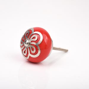 May include: A red ceramic cabinet knob with a silver floral design. The knob is round and has a silver metal stem. The decorative hardware is suitable for drawers, cabinets, and furniture.