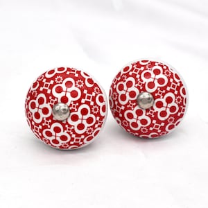 May include: Two round cabinet knobs with a red and white floral pattern. Each knob has a silver-coloured metal screw. The knobs are likely used for drawers or cabinets.