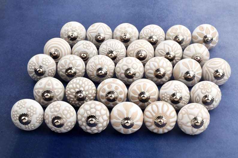 collection of white ceramic drawer knobs with intricate floral patterns and silver accents. The knobs are arranged in a basket lined with peach-colored fabric.