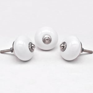 Peut inclure: Trois boutons de meuble en céramique blanche avec une finition brillante. Chaque bouton est orné d'une base décorative argentée et d'une vis filetée pour l'installation. Les boutons sont ronds et présentent un design simple et élégant.