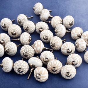 collection of white ceramic drawer knobs with intricate floral patterns and silver accents. The knobs are arranged in a basket lined with peach-colored fabric.