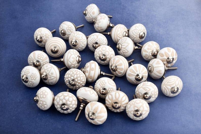 collection of white ceramic drawer knobs with intricate floral patterns and silver accents. The knobs are arranged in a basket lined with peach-colored fabric.