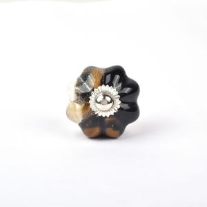 Op de afbeelding: Decoratieve keramische kastknop met een bloemmotief. De knop heeft een mix van kleuren, waaronder zwart, bruin, wit en zilver. Het midden van de knop heeft een zilverkleurig bloemmotief. De knop is ontworpen voor kasten en lades.