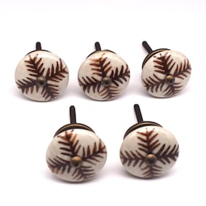 May include: Five white ceramic cabinet knobs with brown fern designs. Each knob has a brass screw.