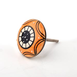 May include: An orange ceramic cabinet knob with a black and white circular design. The knob has a silver threaded stem for installation. The design features black curved lines and white dots on the orange surface. A decorative hardware item.