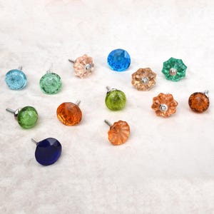Puede incluir: Una colección de doce pomos decorativos para armarios en varios colores, incluyendo azul, verde, naranja y melocotón. Cada pomo presenta una superficie de cristal facetado o agrietado con un tornillo central de metal plateado.