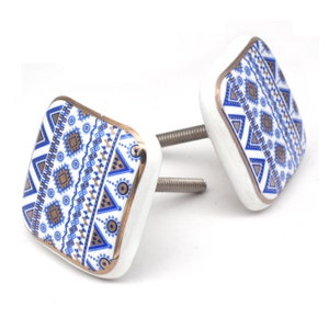 Könnte beinhalten: Zwei quadratische weiße Keramik-Knöpfe mit einem blauen und goldenen geometrischen Muster. Die Knöpfe haben einen goldenen Rand und eine silberne Schraube.