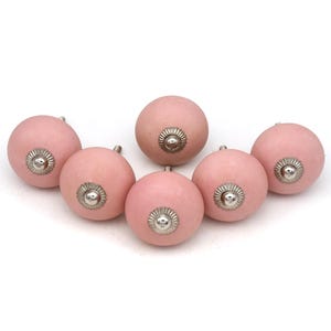 May include: Six pink ceramic cabinet knobs with silver-colored metal accents. The knobs are round with a smooth, matte finish. The metal accents are circular with a detailed, floral-like design. These knobs are suitable for furniture or cabinet upgrades.