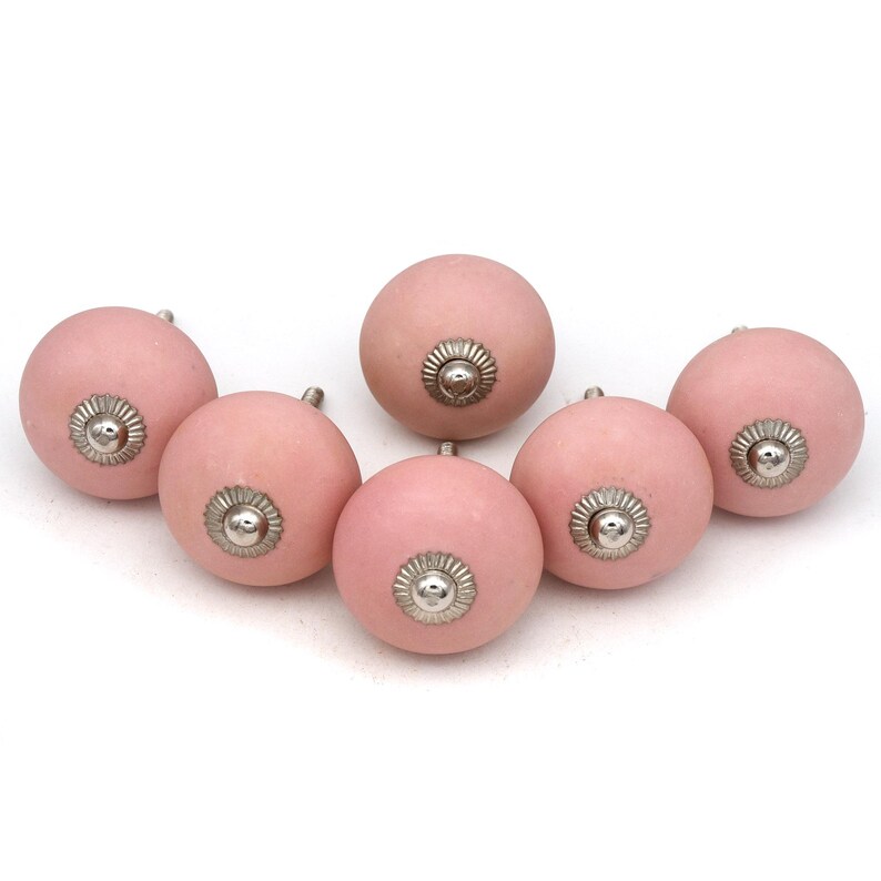 May include: Six pink ceramic cabinet knobs with silver-colored metal accents. The knobs are round with a smooth, matte finish. The metal accents are circular with a detailed, floral-like design. These knobs are suitable for furniture or cabinet upgrades.