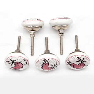 Könnte beinhalten: Set aus fünf weißen Keramik-Schrankknöpfen mit einem rosa Vogeldesign. Jeder Knopf hat eine silberne Metallbasis und Schraube. Die Knöpfe haben ein gepunktetes Muster am Rand und sind für Schubladen oder Schränke geeignet.