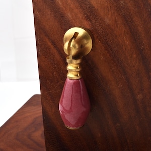 May include: A close-up of a cabinet pull. The pull features a brass mounting plate and a burgundy teardrop-shaped ceramic handle. The handle is attached to the brass hardware with a gold-coloured metal connector. The cabinet is made of dark brown wood.