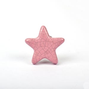 May include: A star-shaped ceramic cabinet knob in a soft pink color. The surface features a distinct dark crackle glaze pattern across the entire front, creating a network of fine lines over the smooth, glossy finish of the star-shaped object.
