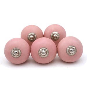 May include: Five pink ceramic cabinet knobs with silver-colored metal accents. The knobs are round with a matte finish and are arranged closely together against a white background. These knobs are suitable for drawers and cabinets.