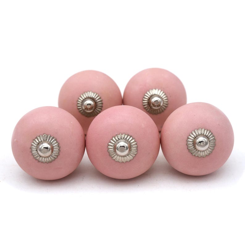 May include: Five pink ceramic cabinet knobs with silver-colored metal accents. The knobs are round with a matte finish and are arranged closely together against a white background. These knobs are suitable for drawers and cabinets.
