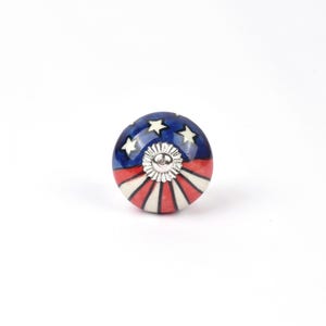 May include: A round cabinet knob with a patriotic design. The knob features a blue background with white stars, red and white stripes, and a silver-colored center. This decorative hardware adds a touch of Americana to any furniture piece.
