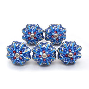 May include: Five blue and white ceramic cabinet knobs with a floral design. Each knob features a silver center and red and yellow accents. The knobs are arranged on a white background.