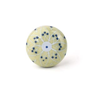 May include: A round cabinet knob with a floral design. The knob is pale yellow with a white floral pattern and navy blue accents. The centre of the flower is white with a navy blue dot.