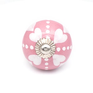 Könnte beinhalten: Ein rosa Keramik-Schrankknopf mit weißem Herzmuster und silberfarbener Basis. Der Knopf ist rund und hat ein dekoratives Blumendesign auf der Basis.