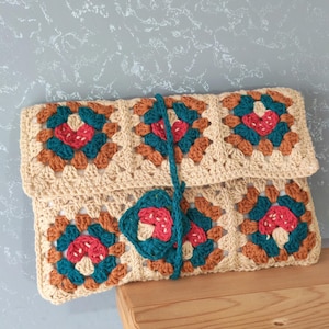 May include: A rectangular crochet clutch bag made of joined granny squares in cream, teal, rust orange, and pink. The bag features a fold-over flap and a long teal crochet strap that wraps around the front to secure the closure with a square motif.