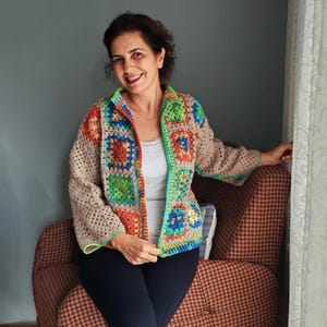 Op de afbeelding: Een kleurrijke, open gehaakte jas. De jas heeft een patchwork-ontwerp met vierkanten in groen, blauw, oranje en rood, omlijst door een beige rand en groene afwerking. De mouwen zijn lang en de jas is open aan de voorkant.