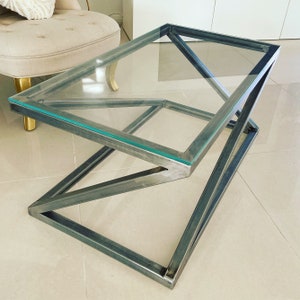 Peut inclure: Une table basse en verre avec un cadre en métal angulaire géométrique. La table a un plateau en verre transparent et un cadre en métal argenté.