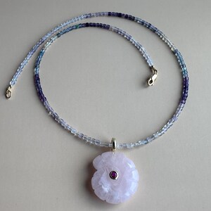 Könnte beinhalten: Eine zarte Halskette mit einem Farbverlauf aus blauen und violetten Perlen. Die Halskette hat einen goldenen Verschluss und einen Anhänger in Form einer hellrosa Blume mit einem kleinen roten Edelstein in der Mitte. Es ist ein Schmuckstück.