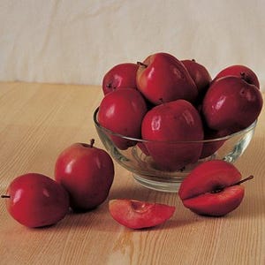 May include: A clear glass bowl filled with several smooth, round, deep red fruits. Two whole fruits sit outside the bowl on the left, while one sliced wedge and one halved fruit with a visible stem are positioned on the right side of the bowl.