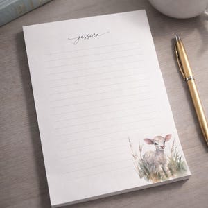 May include: White lined notepad with a watercolor illustration of a lamb in grass. The notepad has the word "Jessica" in script and the text "Resurrection Lamb Notepad". A gold pen and a cup of coffee are next to the notepad.